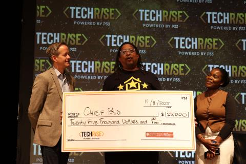 Three people stand on stage in front of a repeating "TechRise, powered by P33" backdrop. In the center, Chief Bud founder Chad King — wearing a black long-sleeve shirt with the yellow Chief Bud house-and-stars logo — speaks while holding an oversized ceremonial check. A man in a tan blazer stands at left and a woman in a brown top and white skirt at right, each steadying one end of the check. The check is dated 11/11/2025, made out to Chief Bud for $25,000, and carries the TechRise and University of Chicago